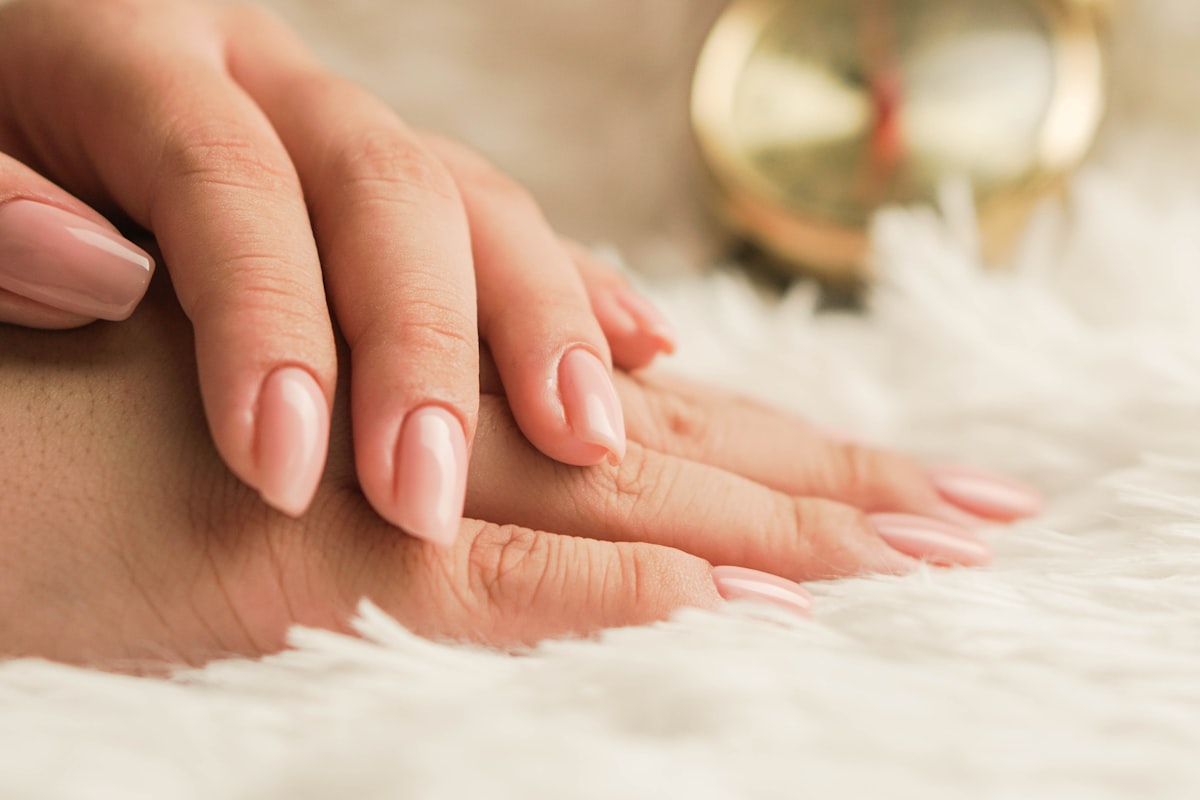 Two hands with a natural nude gel manicure resting on a soft white surface